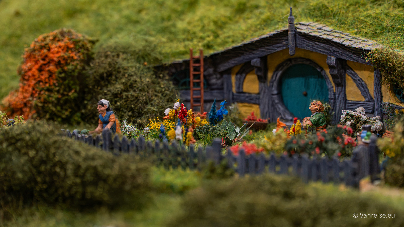 Miniaturhaus mit begrünter Dachfläche, runder blauer Tür, Blumenbeeten und kleinen Figuren.