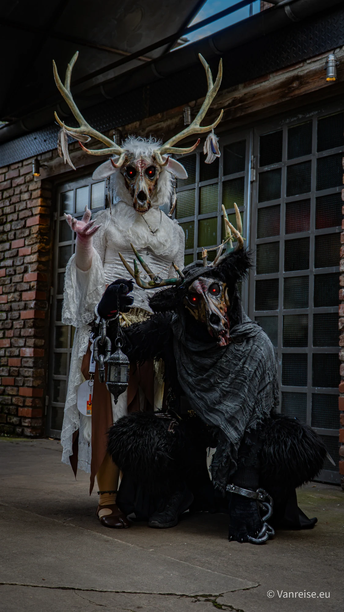 Zwei Horror-Cosplayer mit Geweih- und Schädelmasken vor einer industriellen Hallenfassade.
